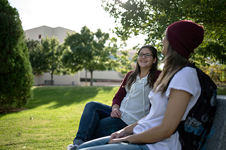 Image of two female students sitting on a bench and talking. In the image background the silhouette of a building can be seen.