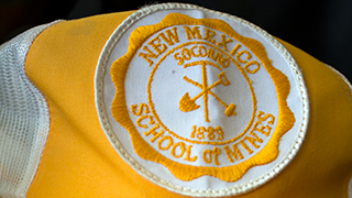 A close up image of a vintage New Mexico Tech baseball cap. The background of the cap is white, and the old New Mexico Tech logo, a shovel and hammer crossed behind a pickaxe standing upright with the word Socorro above the pickaxe to represent the school's location, and "1889" underneath the pickaxe to commemorate the school's founding. "New Mexico School of Mines" circles the logo.  