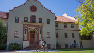 An image of the exterior of the New Mexico Tech gym. The exterior is white stucco in shadows from the sun. A bicyclist is riding in front of the building.