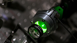 An above view close up image of a laser and lens being used in an experiment. A green glow is seen from the opening behind the lens. A silver circuitboard is seen in the background.
