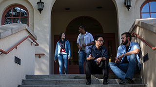 A group of students talking at the entryway to Cramer Hall. Two students are seated on the steps talking and two additional students are talking as they exit the doors.