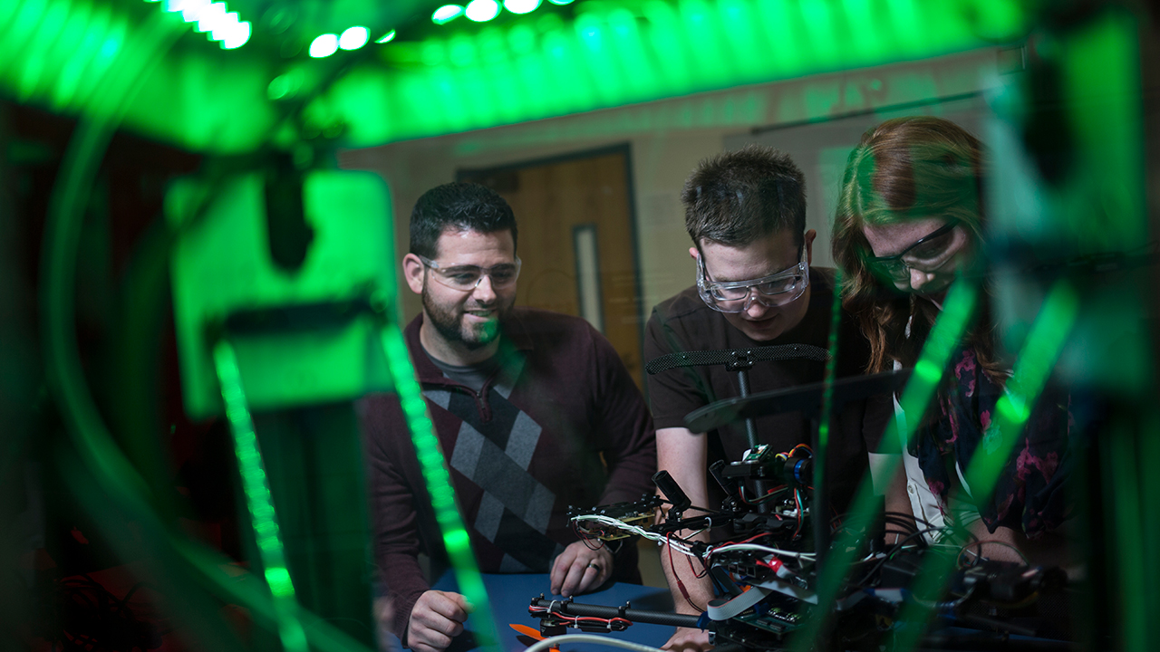 Three students, two male and one female, are working on a drone in an NMT lab space.